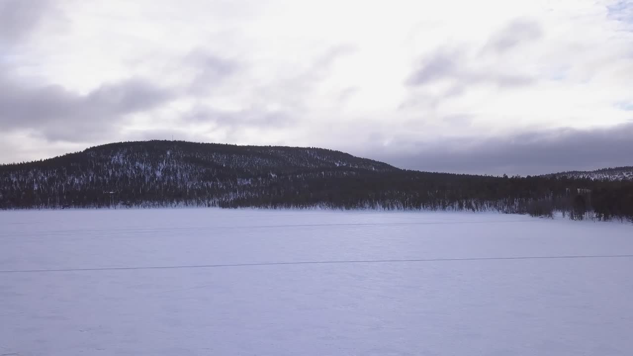 Drone shot flying over and upwards on a frozen lake covered in snow. In the background are hills covered in snow at sunset.