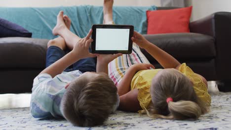 happy caucasian brother and sister lying on floor in living room using tablet, copy space on screen