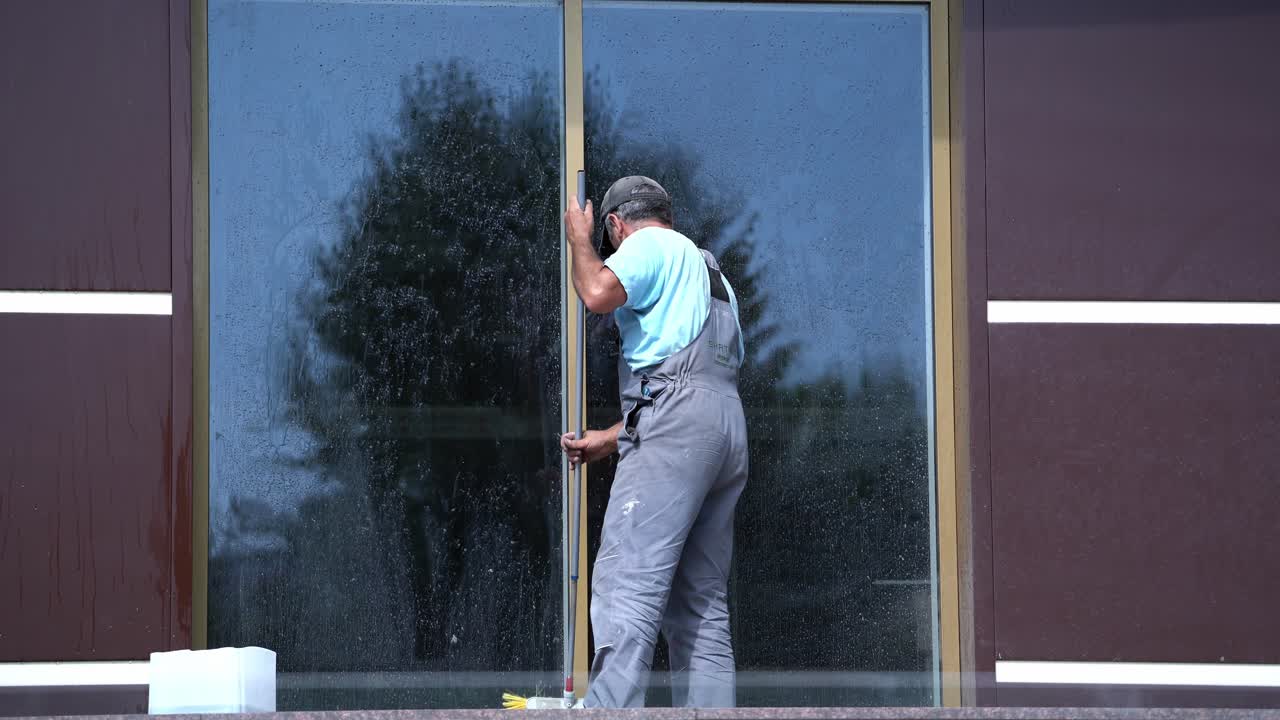 Man washes office window. Back view of a male worker who is cleaning window with a mop outdoors. Washing glass window of office building. Building cleaning services.