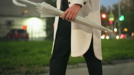 partial view of person swinging folded umbrella during evening stroll with cars passing by, bokeh lights creating glowing atmosphere, urban park path and greenery visible in soft blurred background