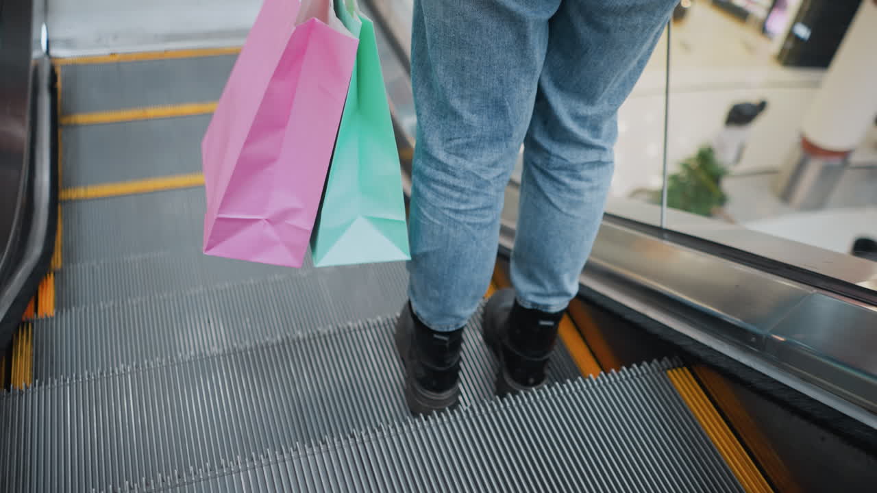 Bagfra set af underben i jeans og sorte støvler, bærende farverige indkøbsposer, træder forsigtigt ud på en rullende rulletrappe i et livligt indkøbscenter