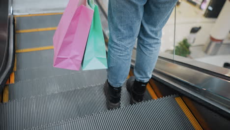 rear view of lower legs in jeans and black boots, carrying colorful shopping bags, stepping out gently on a moving escalator in a lively mall