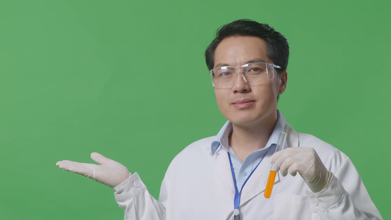 Close Up Of Asian Man Scientist With Orange Liquid In The Test Tube Smiling And Pointing To Side While Standing On The Green Screen Background In The Laboratory