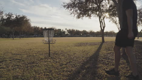 disc golfer throws a disc with a backhand throw next to a disc golf basket into an open field