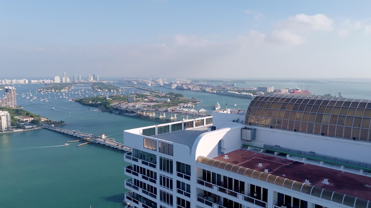 Aerial drone view of Miami’s Biscayne Bay with bridges, luxury yachts, cruise ships, and the port, featuring a rooftop foreground under a partly cloudy sky.
