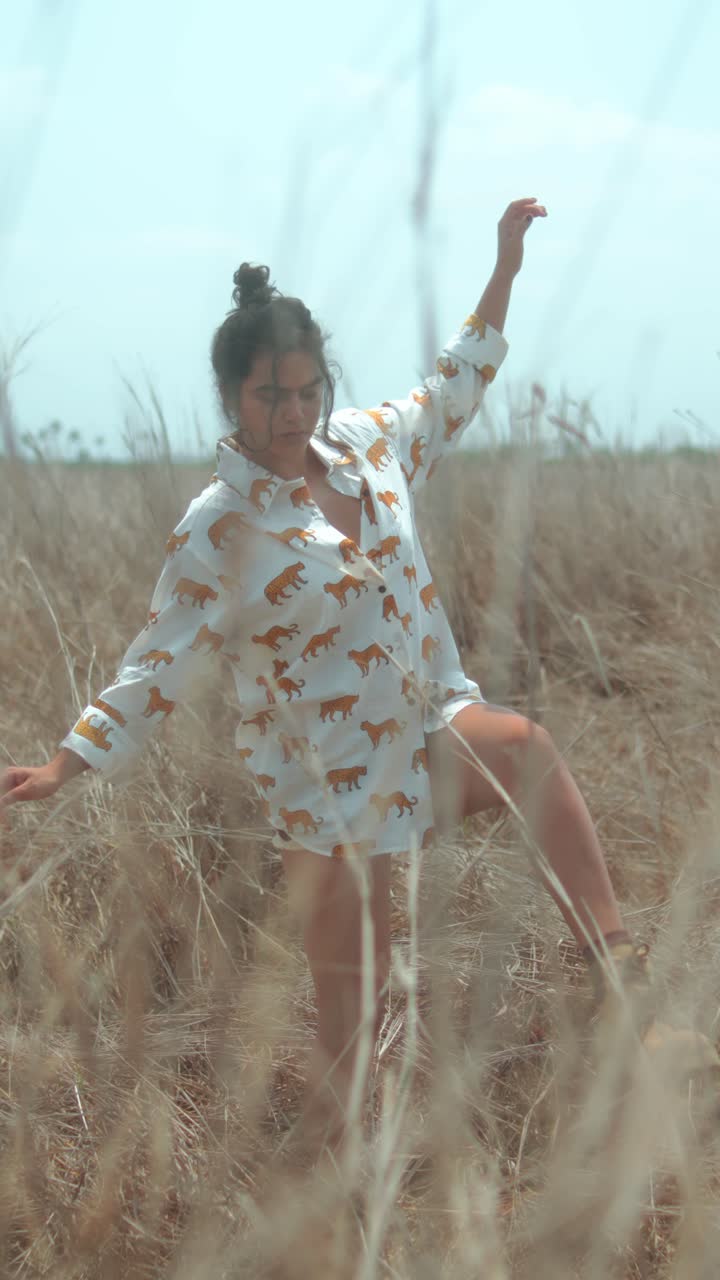 Young woman dancing playfully in nature, surrounded by tall grass, wearing a shirt