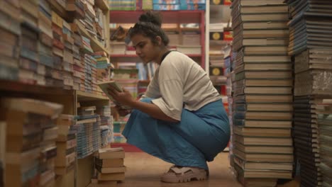 Una-Hermosa-Mujer-Leyendo-La-Portada-Y-El-Título-De-Un-Libro-Mientras-Se-Agacha-En-Medio-De-Una-Isla-En-Una-Librería,-India