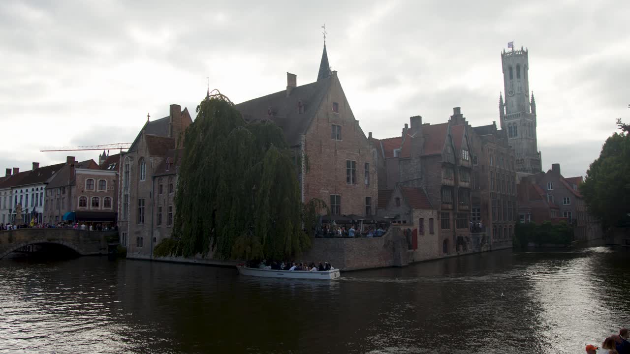 Tour boat glides along canal past medieval architecture, Belfry tower, under overcast natural daylight