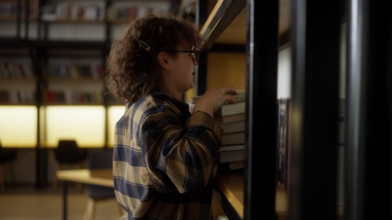 una estudiante con cabello rizado con gafas en una camisa a cuadros pone una pila de libros en un estante en la biblioteca