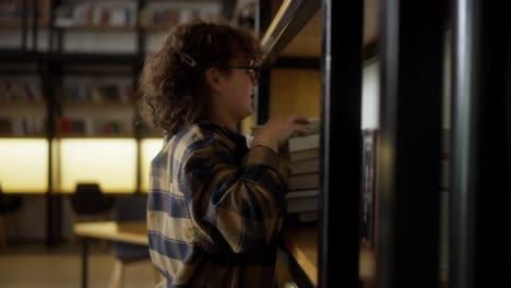 a girl student with curly hair with glasses in a checkered shirt puts a stack of books on a shelf in the library