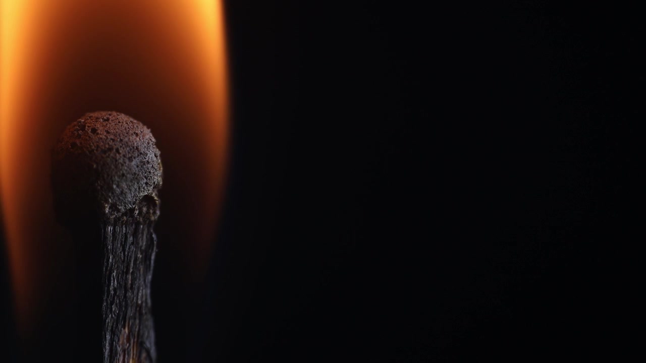 A lit wooden matchstick burns with a bright yellow-orange flame against a deep black background, creating strong contrast and dramatic lighting.