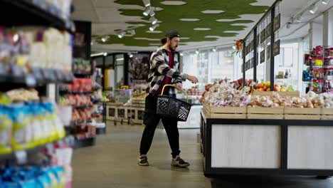 Vista-Lateral-De-Un-Hombre-Confiado-Con-Una-Camisa-A-Cuadros-Y-Una-Gorra-Caminando-Por-El-Supermercado-Y-Colocando-Los-Productos-Que-Necesita-En-Una-Canasta-Durante-Sus-Compras.