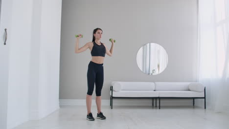 levantar las pesas sobre la cabeza realizando ejercicios para los hombros. entrenamiento en casa en el apartamento. una mujer morena con cabello largo y una hermosa figura