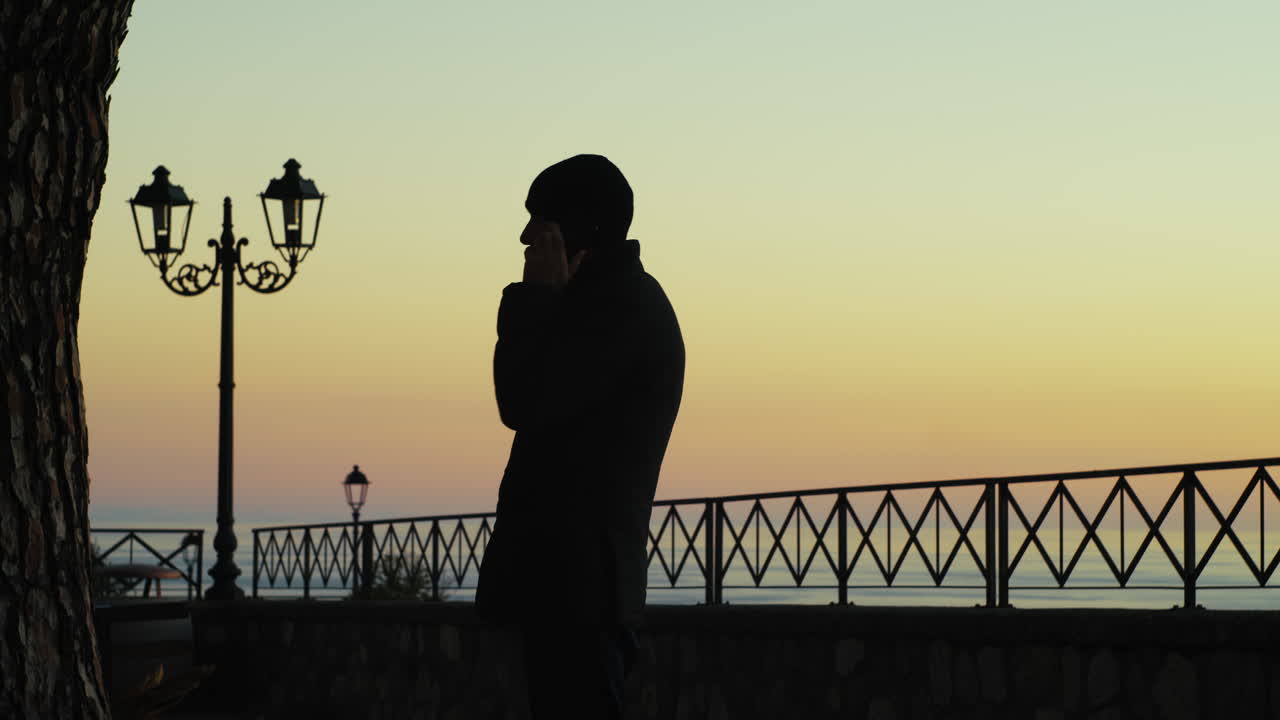 발코니에서 해가 지는 동안 전화로 이야기하는 익명의 남자의 실루