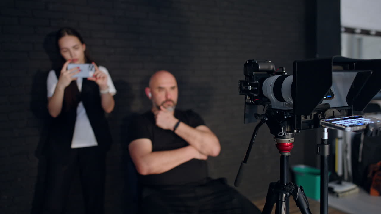 Modern professional camera on tripod recording a video. Cameraman sits on the chair and girl taking video on her phone in blur.