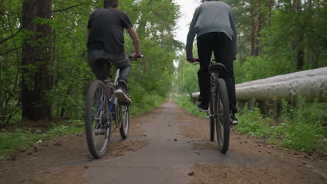 two brothers riding bicycles along a paved untarred path surrounded by lush green trees, one of them stands while cycling, while a log of wood lies along the side of the road