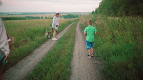 back view of people walking home along a dirt path through lush greenery, a woman in a hat walks with a dog on a leash, while a young boy in a green shirt follows behind