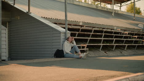 young lady sits thoughtfully on stadium walkway, resting her head on her hand, leaning on a pole with her black bag beside her, she gazes upward, immersed in quiet reflection