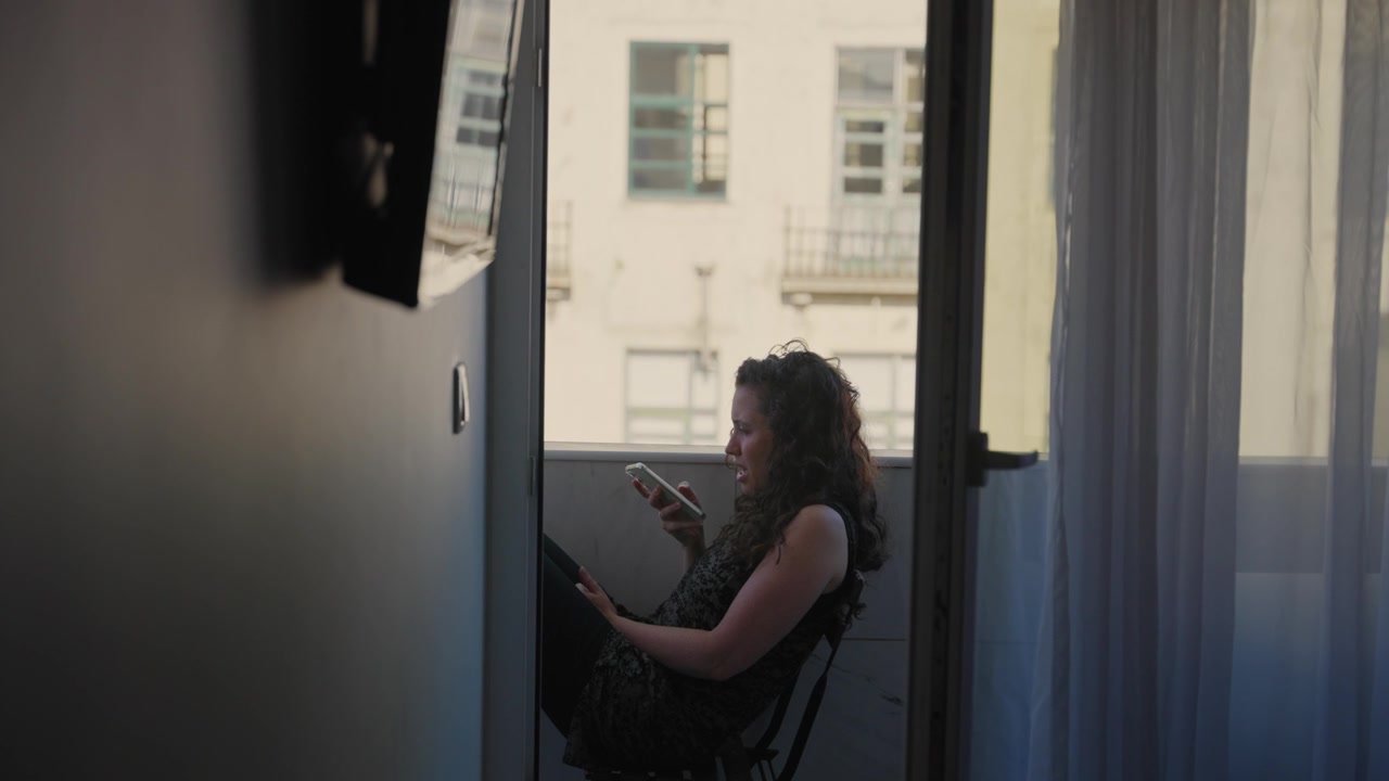 Giovane donna seduta su un piccolo balcone europeo nel suo appartamento che registra un messaggio audio sul telefono cellulare. Giorno d'estate, come turista o abitante del luogo che si gode l'ombra parlando con amici o familiari al telefono.