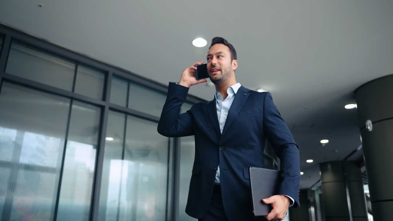 Um empresário profissional ocupado caminhando no corredor de um escritório moderno enquanto realiza várias tarefas com um celular e um tablet digital em um ângulo baixo de rastreamento