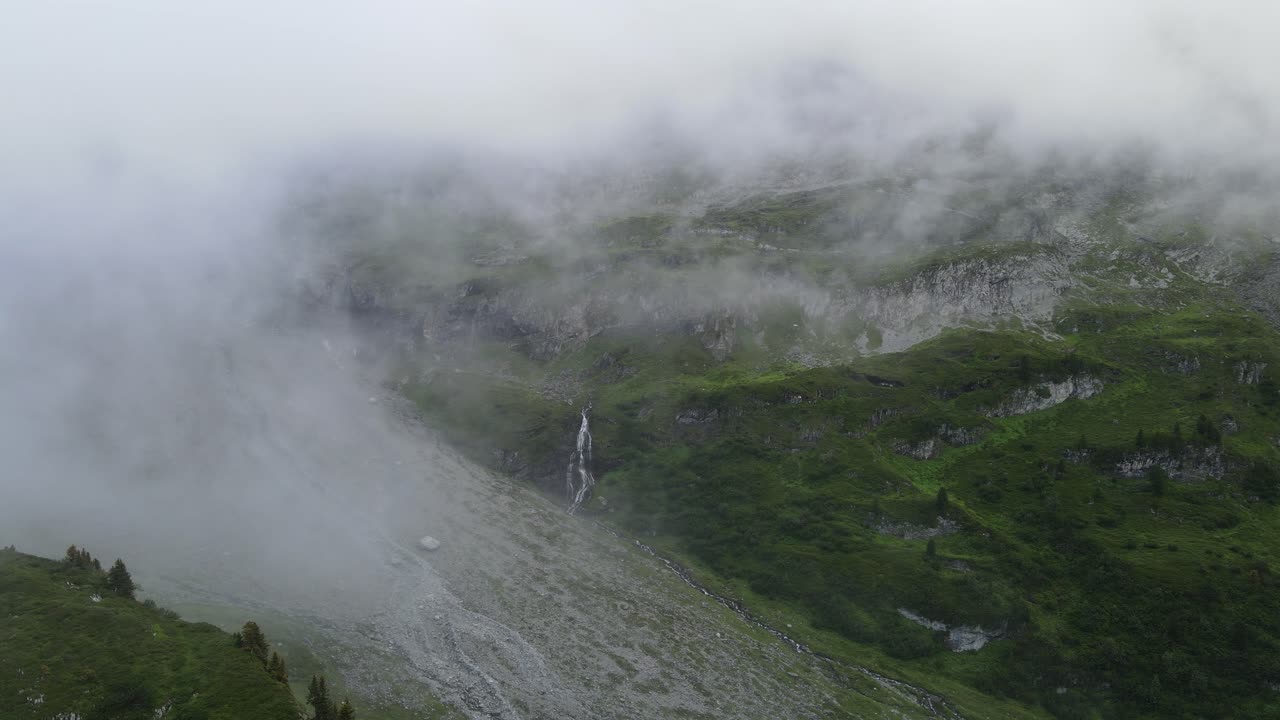 Alpský vodopád Engstlenfall poblíž jezera Engstlensee ve Švýcarsku. Mlhavý vodopád kaskádovitě padá po skalnatých svazích podél Via Alpina, obklopený mraky, zeleným alpským terénem a odlehlou horskou scenérií.