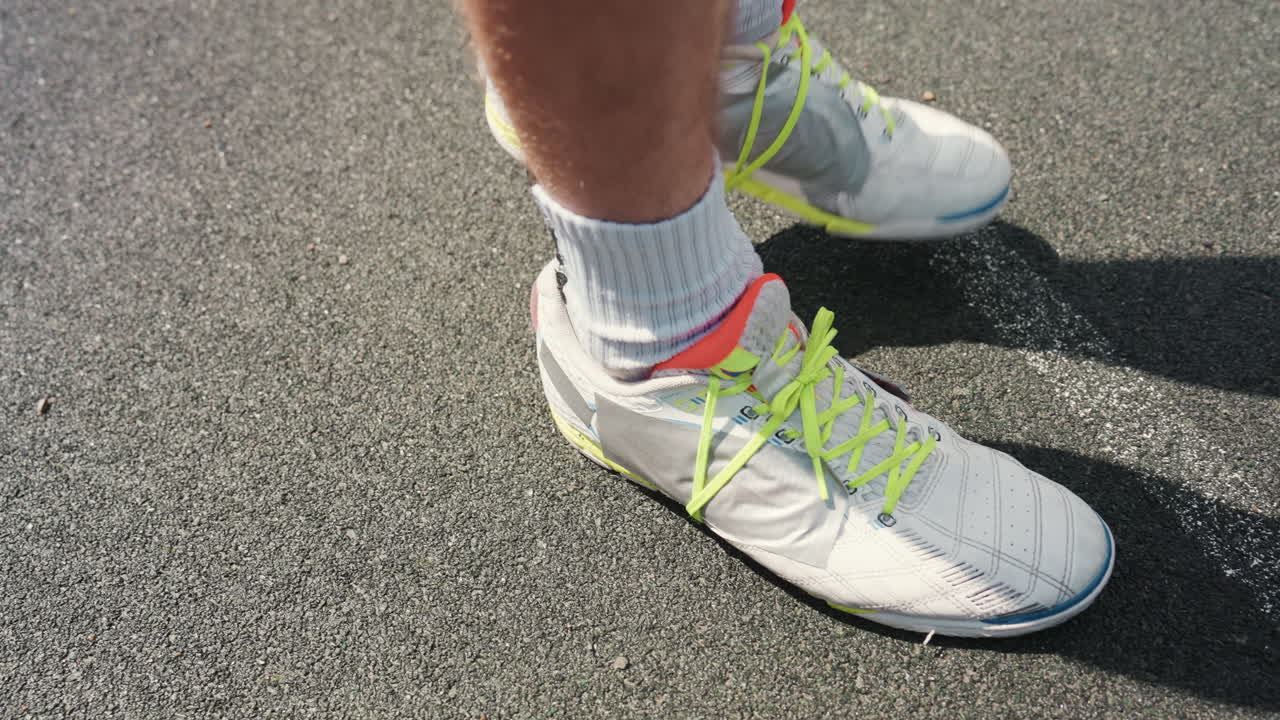 Un atleta si aggiusta le scarpe da corsa bianche sulla pista, lacci neon e calzini sportivi sulla corsia di asfalto, primi piani dei piedi e dell'ombra, posizione concentrata e rituale pre-partenza che segnala la preparazione per lo sprint o la sessione di addestramento