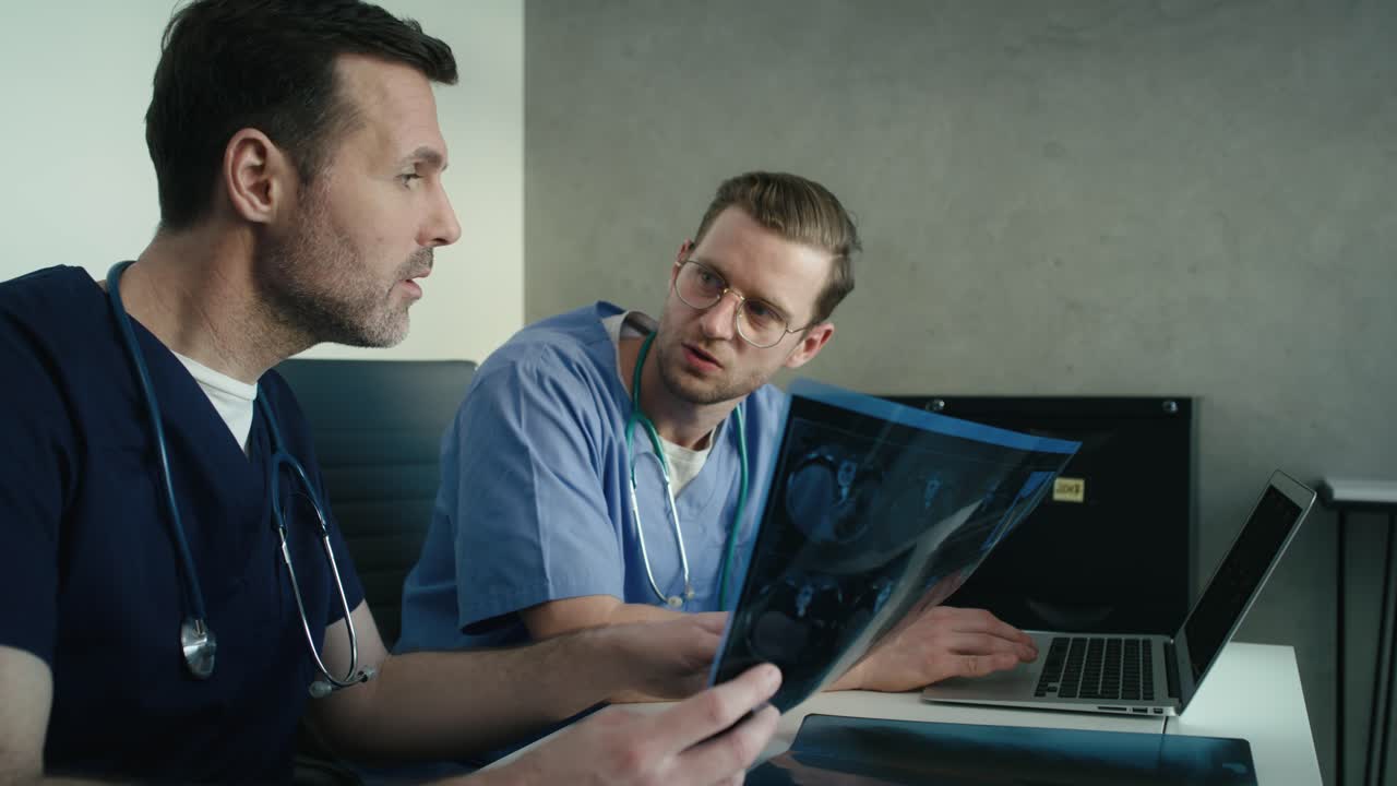 Vista laterale di due medici caucasici maschi che discutono una radiografia medica su una lavagna e un laptop.