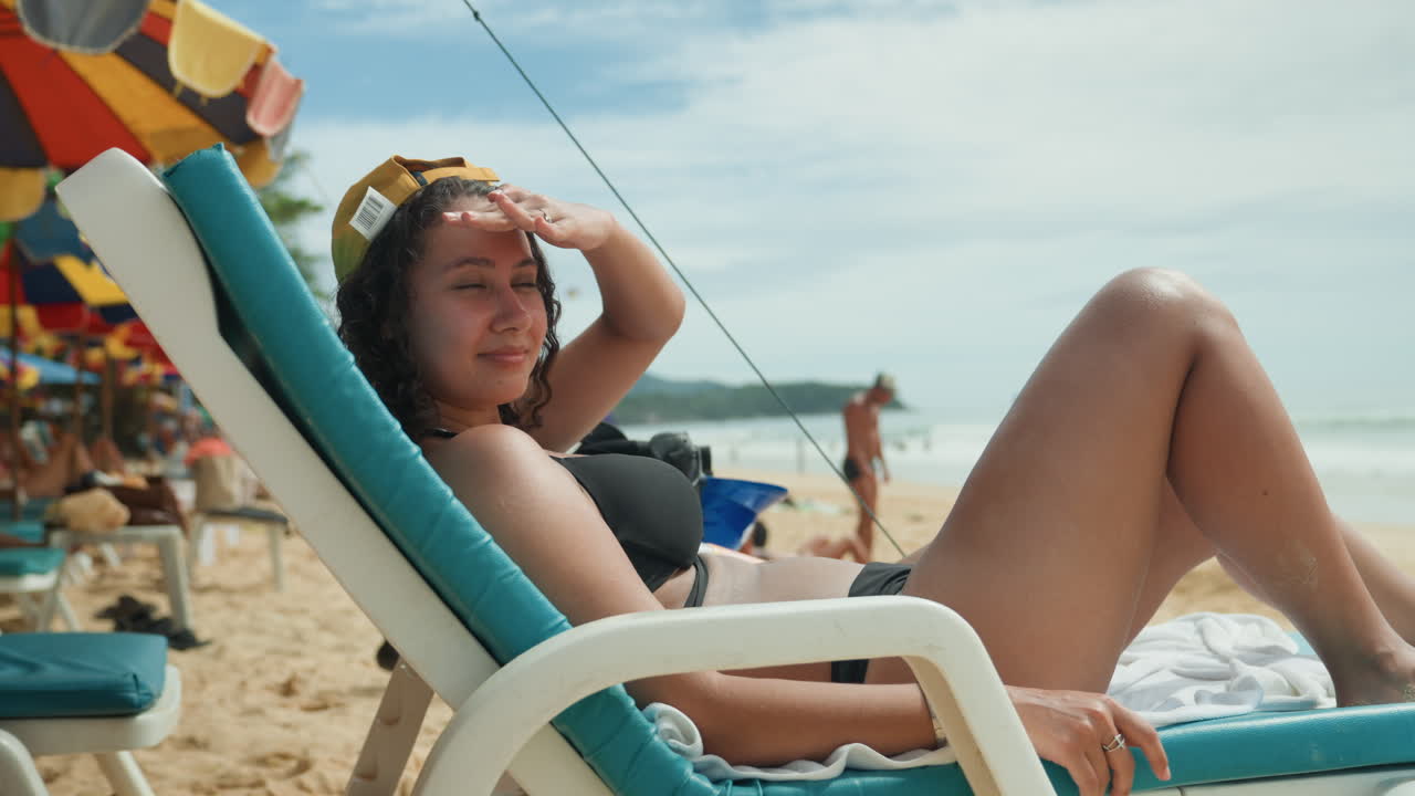 Young woman lounging on beach chair under striped umbrella, adjusting sunglasses, soaking sun, relaxed posture, distant swimmers and waves, warm light, resort atmosphere, carefree summer mood