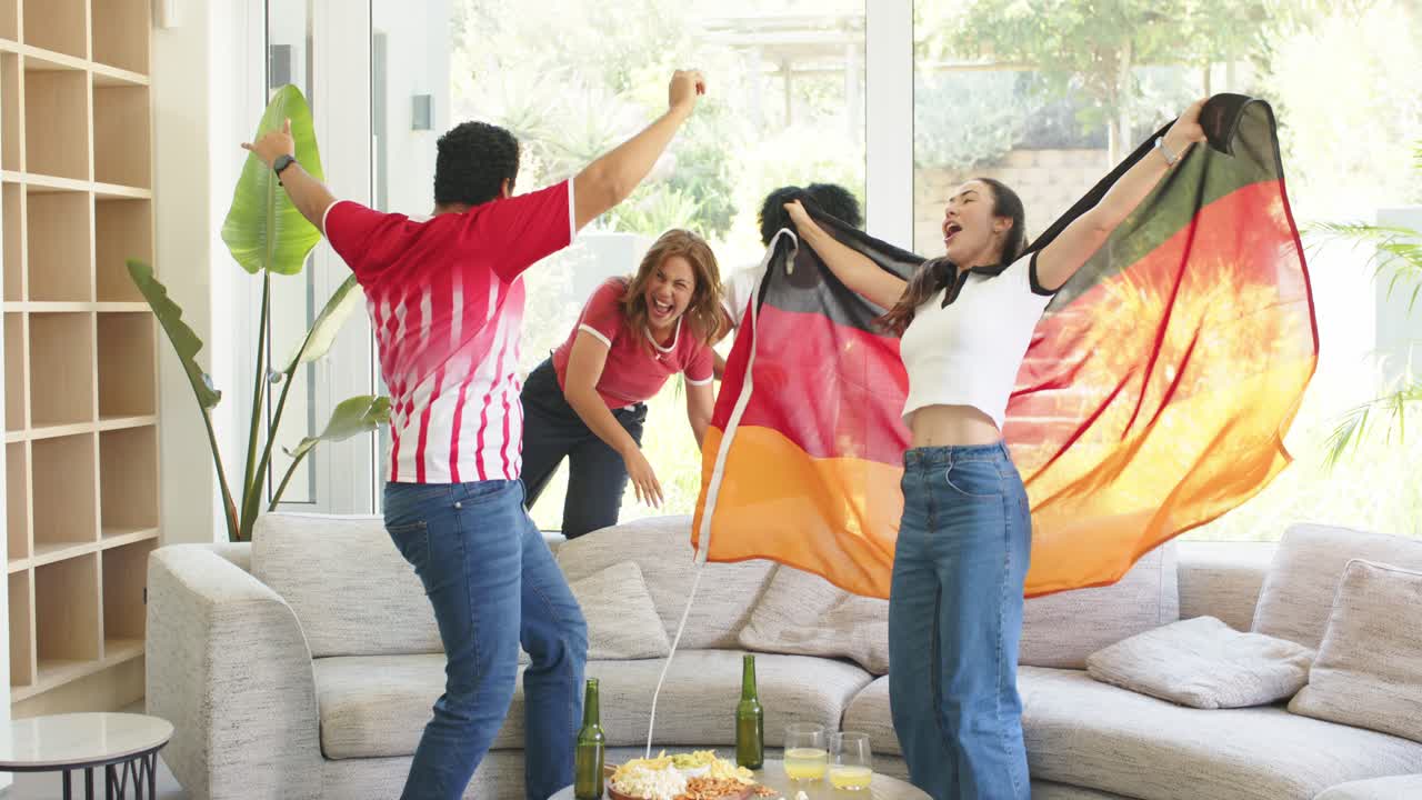 Grupo diverso reagindo a um jogo na sala de estar, torcedor com camisa dançando enquanto mulher acena bandeira tricolor.