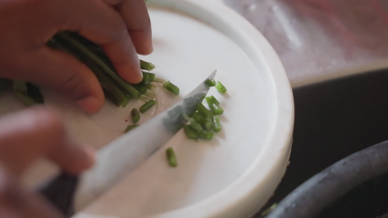 Mani scure che tritano velocemente erbe fresche. Un apprendista chef trita basilico e prezzemolo su un tagliere bianco con un rapido movimento del coltello, formando una piccola guarnizione a cubetti, preparazione concentrata in una cucina urbana, precisa.