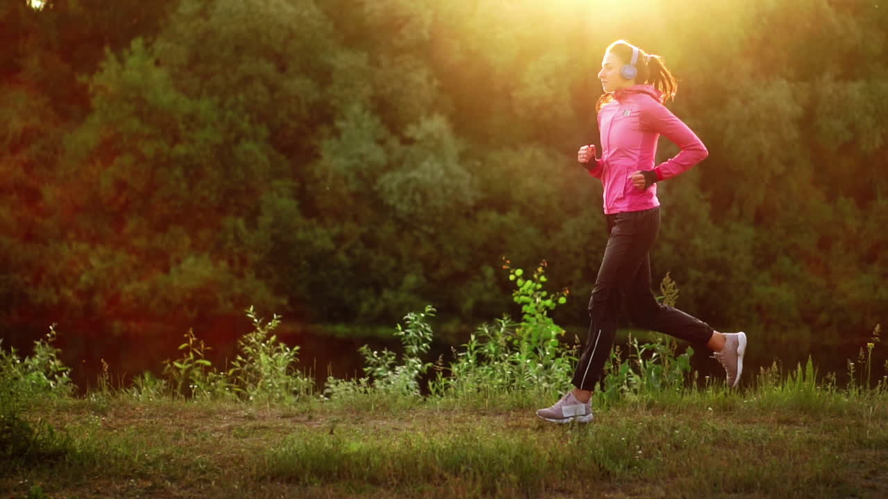 Brunette with long hair in headphones runs along the river in the Park in the morning at sunrise in the summer in a pink jacket and black pants