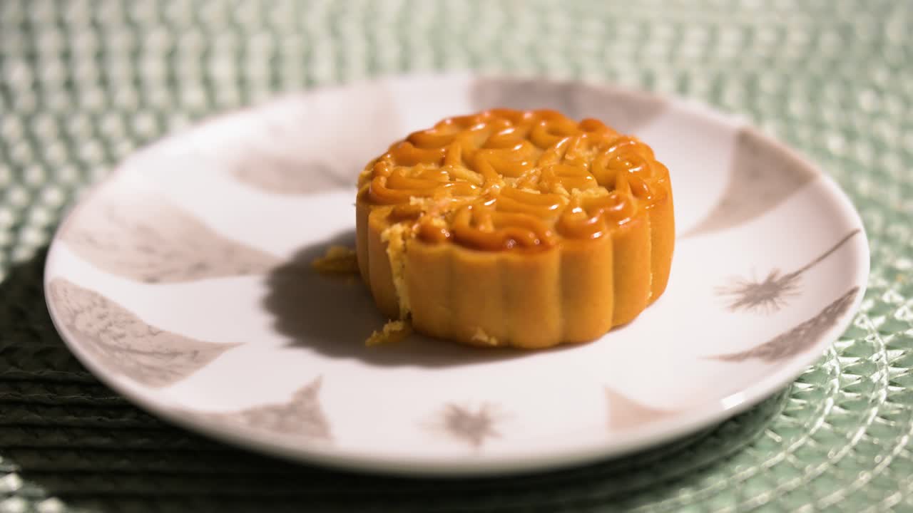 A close-up shot of a golden-brown Chinese mooncake on a plate. The pastry, featuring an intricate pattern, is a traditional dessert eaten during the Mid-Autumn Festival