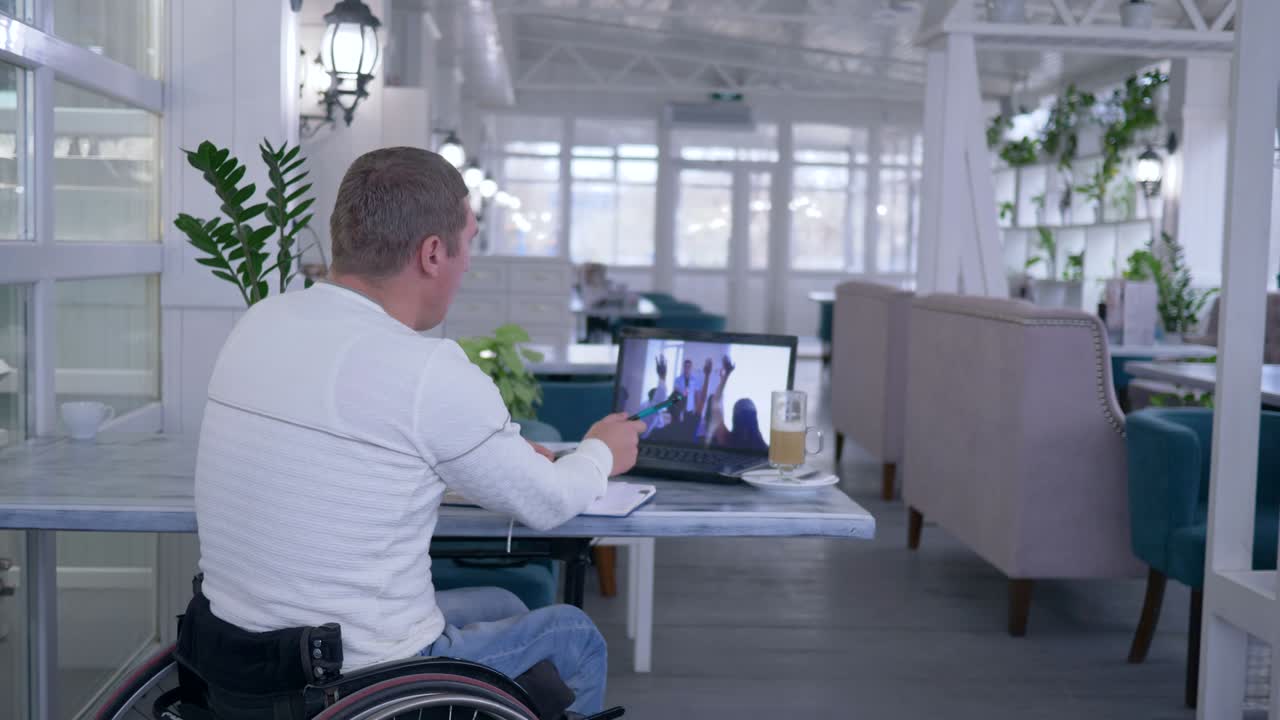 istruzione per corrispondenza, allievo uomo disabile in sedia a rotelle scrive appunti in quaderno e guarda formazione online su computer portatile seduto a tavolo in un ristorante