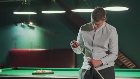 game man in white shirt and grey trousers sits on green pool table, holding cue stick and carefully applying red cover with focused expression as camera zooms in. background features billiards hall