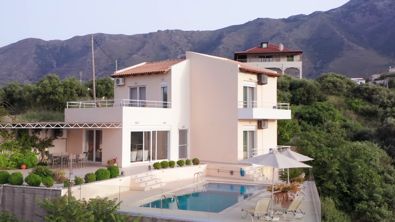 antena baja en sentido contrario a las agujas del reloj vuela alrededor de una lujosa villa griega con piscina con montañas en el fondo y follaje verde en primer plano