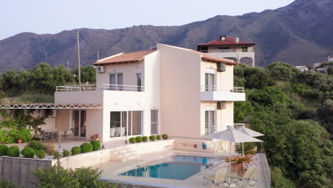 Low-Aerial-Counter-Clockwise-Fly-Around-of-Luxury-Greek-Villa-with-Pool-with-Mountains-in-Background-and-Green-Foliage-in-Foreground