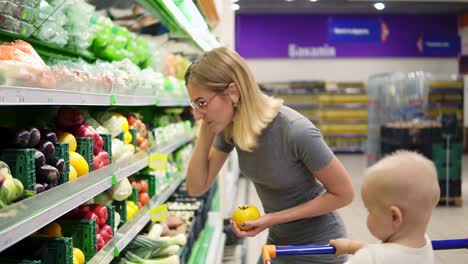 Die-Junge-Mutter-Wählt-Gelbe-Tomaten-Und-Anderes-Gemüse-Aus,-Während-Ihr-Kleines-Kind-In-Einem-Einkaufswagen-Sitzt.-Familie