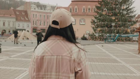 Vista-Trasera-De-Una-Mujer-Hipster-Con-Gorra-Caminando-En-La-Plaza-Del-Consejo-Durante-La-Temporada-Navideña-En-Brasov,-Rumania