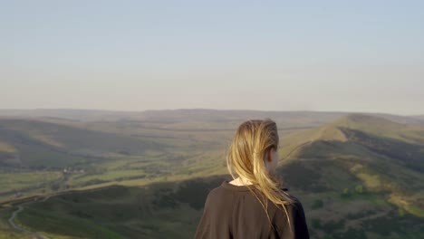 stabilised shot of young blonde woman with hair over one shoulder on top of mam tor, castleton, peak district, england admiring the view of green rolling hills and blue skies