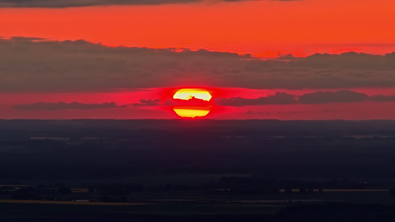 Dramatic aerial descending close-up of massive glowing red and yellow setting sun disk split by dark cloud band against intensely fiery red-orange sky above dark flatlands