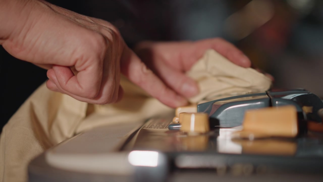 Closeup hands cleaning recorder knobs with cloth, conservator wipes beige rag across yellow controls under soft studio light, tender strokes remove dust during careful preservation routine