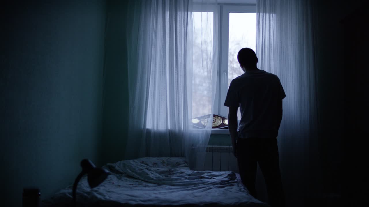 어두운 침실의 창밖을 바라보는 남자