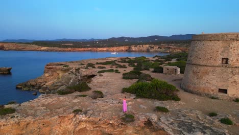 carefree woman in pink dress with open arms enjoying freedom and breathtaking view on ibiza island near torre des savinar at sunset. great aerial view flight overflight flyover reverse drone