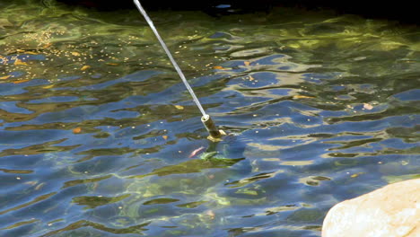 Metal-nozzle-spraying-continuous-stream-of-water-within-a-park-fountain-pond-with-leaves-floating-nearby