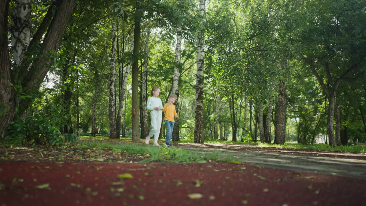 Young girl and boy walk side by side along forested park path covered in shade and sunlight, enjoying peaceful nature together as siblings bond during relaxing outdoor stroll in lush natural scenery