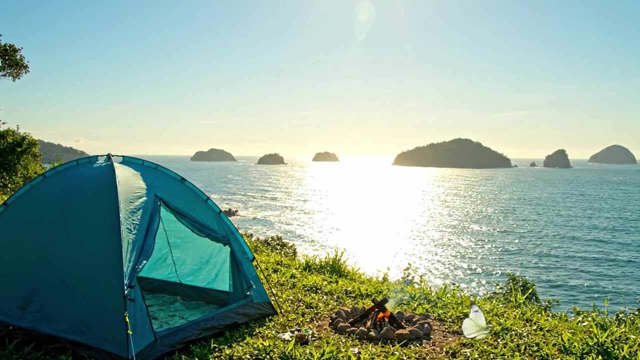 テントと焚き火があり、きらめく海と遠くの島々が広がる息をのむような景色を望む、澄んだ空の下の穏やかな海岸線での静かなキャンプ体験