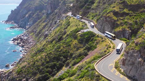 Una-Toma-Aérea-De-Un-Convoy-De-Autobuses-Que-Viajan-Por-Una-Peligrosa-Carretera-De-Montaña-Estrecha-A-Lo-Largo-Del-Océano-Chapmans-Peak-Road,-Cerca-De-Ciudad-Del-Cabo,-Sudáfrica-1