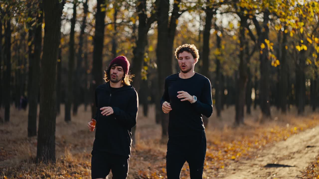 Front view of two brunette guys in black sportswear running and stopping during their morning jog in a sunny autumn park in the morning