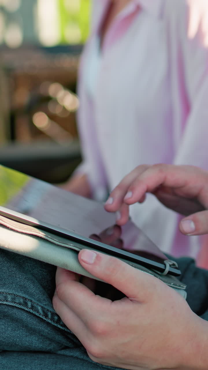 mujer experta en tecnología en rosas multitarea, operando una tableta mientras escribe en su portátil, acompañada por un hombre enfocado escribiendo a su lado, en un telón de fondo arquitectónico borroso