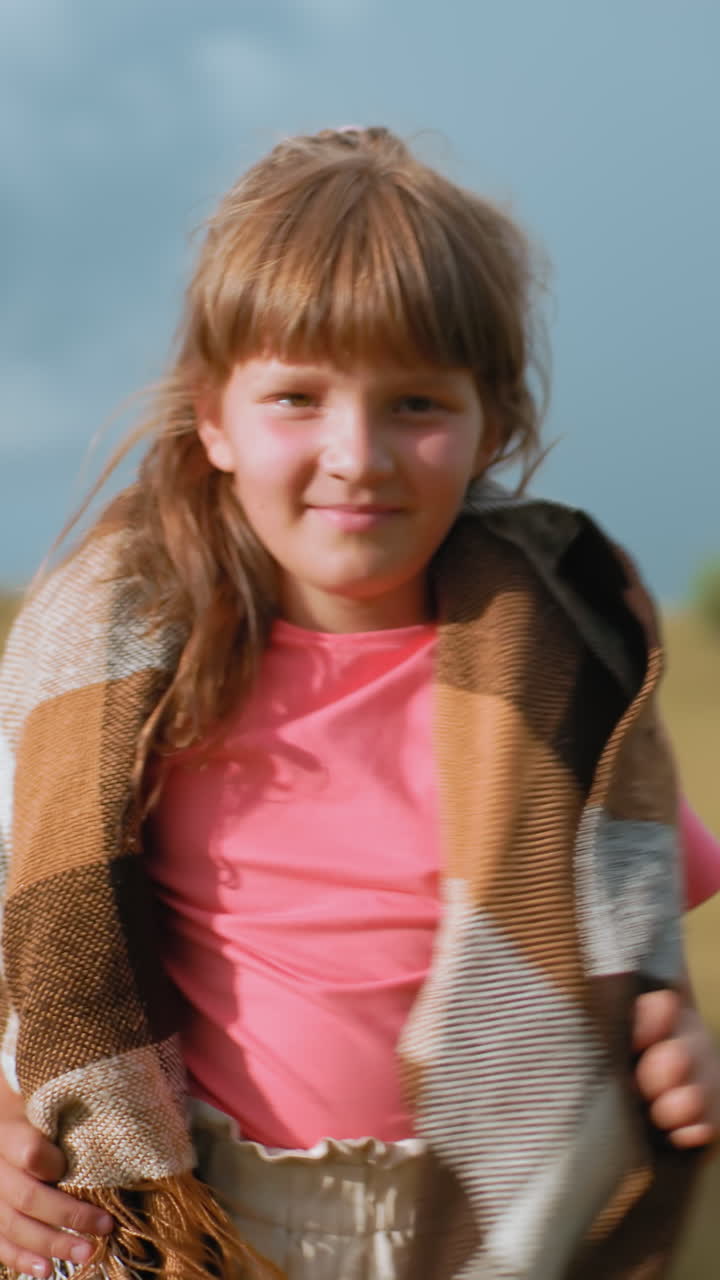 una niña confiada con una camiseta rosa caminando con gracia a través de un vasto campo abierto con una manta sobre los hombros, el viento soplando, balas de heno doradas en la distancia